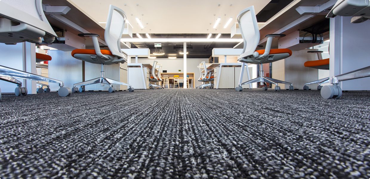 Low-angle view of a modern office space with gray carpet, white desks, and ergonomic chairs, emphasizing a long corridor-like perspective.